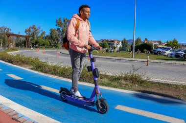 Genç Latin bir adam mavi bisiklet yolunda bir e-scooterla parktan çıkıyor. Motorlu scooter 'la şehirden işe gidip geliyor. Boşluğu kopyala, arkaplan.
