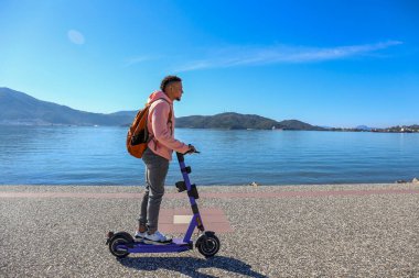 Kıyı şeridi boyunca kiralık bir e-scooter 'la adaların güzel manzarası olan İspanyol genç bir adam. Boşluğu kopyala, deniz manzarası arkaplanı.