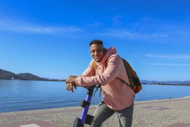 Kıyı şeridi boyunca kiralık bir e-scooter 'la adaların güzel manzarası olan İspanyol genç bir adam. Boşluğu kopyala, deniz manzarası arkaplanı.