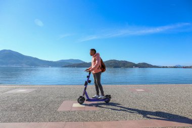 Kıyı şeridi boyunca kiralık bir e-scooter 'la adaların güzel manzarası olan İspanyol genç bir adam. Boşluğu kopyala, deniz manzarası arkaplanı.