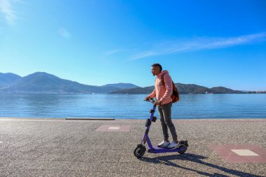 Kıyı şeridi boyunca kiralık bir e-scooter 'la adaların güzel manzarası olan İspanyol genç bir adam. Boşluğu kopyala, deniz manzarası arkaplanı.