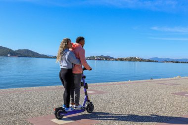 Adaların güzel manzarası ile gezinti alanında birlikte kiralık bir e-scooter süren genç Latin çift. Boşluğu kopyala,