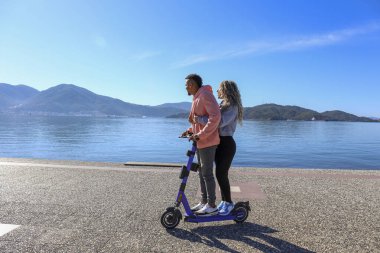 Adaların güzel manzarası ile gezinti alanında birlikte kiralık bir e-scooter süren genç Latin çift. Boşluğu kopyala,