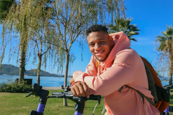Kiralık bir e-scooter 'ın gidonuna yaslanan ve gülümseyen genç bir İspanyol. Parktaki motorlu mutlu latin adam. Kapat, kopyala alanı, yeşillik arkaplanı.