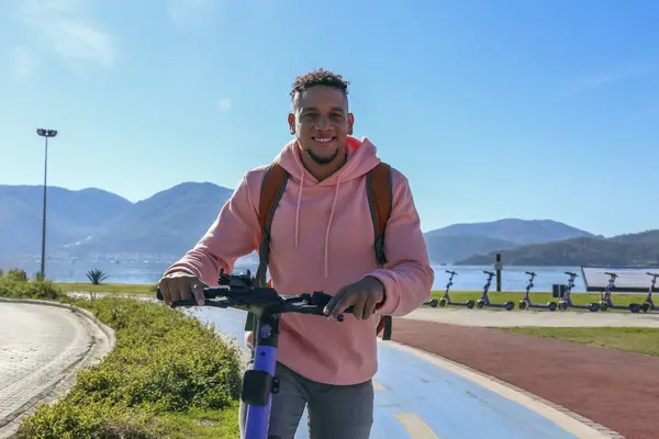 Güzel ada manzaralı mavi bir bisiklet yolunda e-scooter süren genç bir latin. Motorlu scooter 'la şehirden işe gidip geliyor. Boşluğu kopyala, tropikal hedef arkaplan.