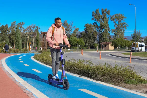 Genç Latin bir adam mavi bisiklet yolunda bir e-scooterla parktan çıkıyor. Motorlu scooter 'la şehirden işe gidip geliyor. Boşluğu kopyala, arkaplan.