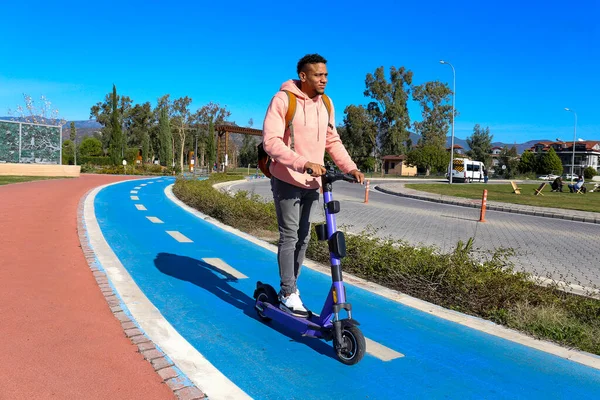 Genç Latin bir adam mavi bisiklet yolunda bir e-scooterla parktan çıkıyor. Motorlu scooter 'la şehirden işe gidip geliyor. Boşluğu kopyala, arkaplan.