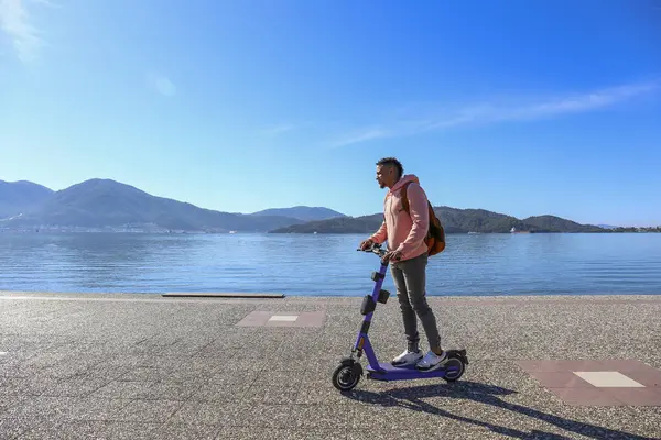 Kıyı şeridi boyunca kiralık bir e-scooter 'la adaların güzel manzarası olan İspanyol genç bir adam. Boşluğu kopyala, deniz manzarası arkaplanı.