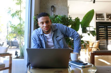 Latin kökenli genç bir adam, elinde laptopuyla kafeteryada oturuyor ve çalışıyor. uzak iş konsepti. Kapat, kopyala boşluk, arkaplan.