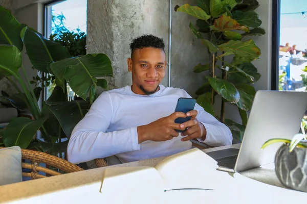Young latin guy with a phone and laptop working remotely from a coffee shop. Hispanic man texting and smiling. Close up, copy space, background.