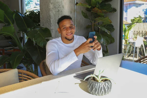 Young latin guy with a phone and laptop working remotely from a coffee shop. Hispanic man texting and smiling. Close up, copy space, background.