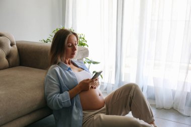 Hamilelik izleme uygulaması konsepti. Genç hamile bir kadın yerde oturuyor ve telefonunu kontrol ediyor. Kapat, kopyala boşluk, arkaplan.