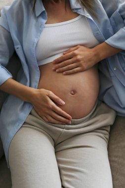 Hamileliğinin üçüncü üç ayında olan genç bir kadın. Kolları karnında, koltukta oturan hamile bir kadın. Çocuk konsepti bekliyorum. Arkaplan, boşluğu kopyala.