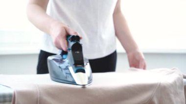 Ütü masasında açık pembe kapüşonlu kadın ütülüyor. Demirden sıcak kökler. Elektrikli ütü tutan kadın, ev işleri. Rutin ev işleri. Yüksek kalite 4k görüntü