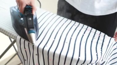 Ütü masasındaki çizgili elbiseleri ütüleyen kadın. Kıyafetlerin üzerine sıcak ütü bırak. Demirden sıcak kökler. Elektrikli ütü tutan kadın, ev işleri. Yüksek kalite 4k görüntü