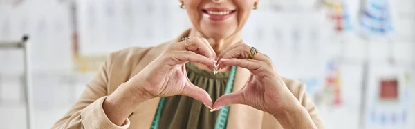 Happy middle aged lady fashion designer with tape measure shows heart with hands standing on blurred background in spacious studio closeup