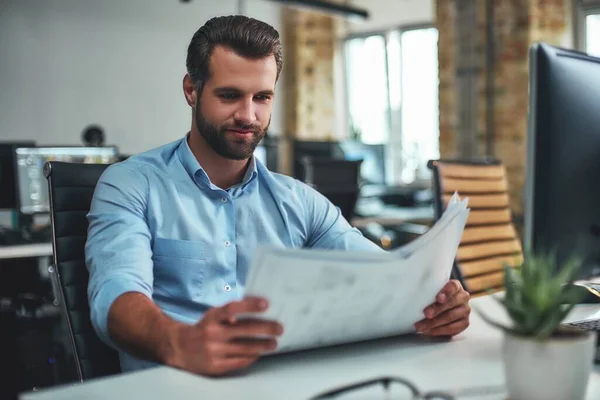 Great work Concentrated bearded engineer in formal wear looking at blueprint and smiling while sitting in the modern office. Architecture concept