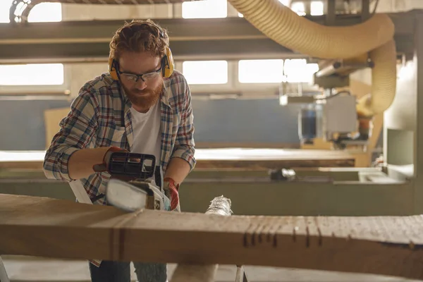 Professional carpenter in glasses is cutting wood with chainsaw on carpentry workshop