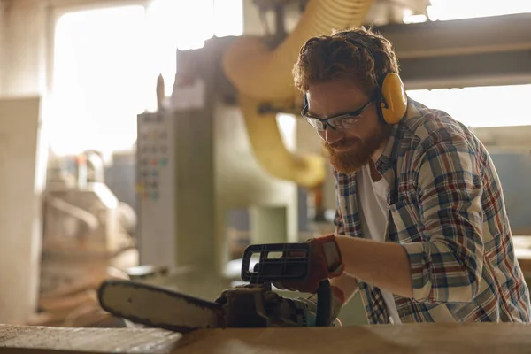 Woodworker in glasses is cutting wood with chainsaw on carpentry workshop. High quality photo