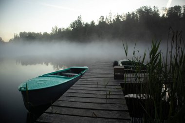 Misty Serenity: Teal Kürek Teknesi ve Tahta Ayak Köprüsü Kuzey Avrupa 'da Yaz Sabahı Sisi tarafından Kucaklandı
