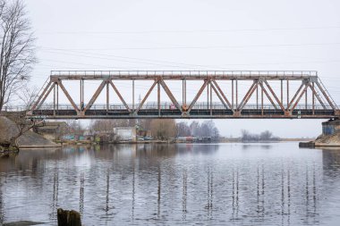 Bulutlu bahar gününde nehrin üzerindeki metal bir köprü. Riga, Letonya 'da doğal ilkbahar manzarası.