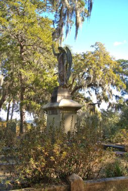 Huge stone statue in historic Bonaventure Cemetery with spanish moss hanging from live oaks over the grave sites in the early morning sunlight