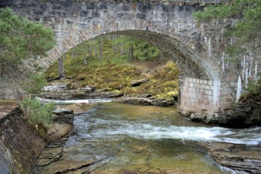 Altında renkli İskoç akıntısı olan taş kemer köprüsü baharın başlarında bulutlu gökyüzü ve yağmurlu