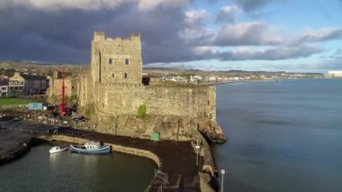 Ortaçağ Norman Kalesi ve Carrickfergus Limanı. Gün batımında Belfast yakınlarında. Su yansıması, marina, tekne rampası, dalgakıran ve kasabalı 4K uçuş videosu. Fırtınalı bulutlu dramatik gökyüzü 