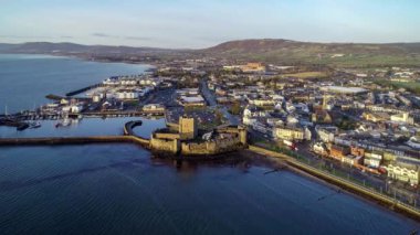 Belfast Lough, Carrickfergus 'taki Ortaçağ Norman Kalesi' nde gün doğumunda Belfast yakınlarında. Limanı, marinası, dalgakıranı, yatları, otoparkı, kasabası ve arka planda Belfast manzarası olan 4K havaalanına yaklaşıyoruz. Kış, gün doğumu.