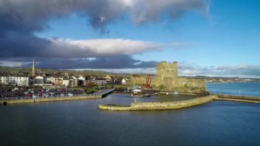 Ortaçağ Norman Kalesi ve Carrickfergus Limanı. Gün batımında Belfast yakınlarında. Marina, tekne rampası, dalgakıran ve kasabalı Aerial 4K uçuş / yaklaşma / açığa çıkarma videosu. Mavi gökyüzü ve kara fırtınalı bulutlar. Gün batımı ışığı 