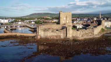 Kuzey İrlanda, Belfast yakınlarındaki Carrickfergus 'taki Ortaçağ Norman Kalesi' nde gün doğumunda. Su yansımalı, marinalı, kasıklı, dalgalı 4K uçuş videosu. Arka planda Belfast 'ın uzak görünümü
