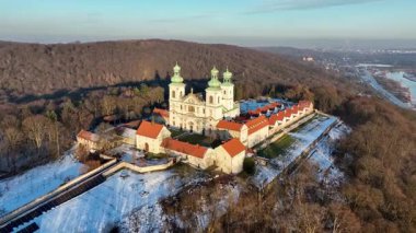 Bielany, Krakow, Polonya 'daki tepedeki ormanda Camaldolese manastırı ve barok kilisesi, kışın günbatımında 4K hava klibi. Arka planda Vistula Nehri ve Krakow şehrinin uzak manzarası