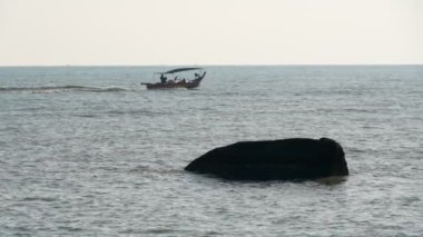 Ön planda belirgin bir kayayla ağır çekim teknesi okyanusta hareket ediyor.