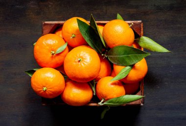Olgun mandalinalar yeşil yapraklarla dolu açık mavi ahşap bir masa üzerinde ahşap bir sandık, meyvenin tazeliğini sergiliyor.
