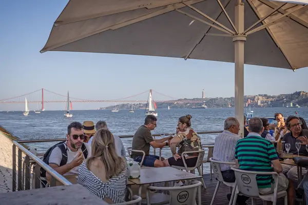 12 Ekim 2023 'te Lizbon, Portekiz' de Tagus Nehri 'nin kıyısında açık havada yenen restoranlar ve ikonik Altın Kapı stili 25 Nisan köprüsü arka planda.