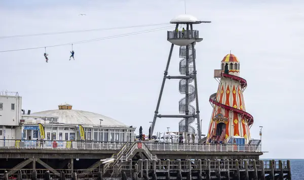stock image Pair of intrepid youngsters taking selfies on zip wire on Bournemouth pier with colourful helter skelter in background at Bournemouth Beach, Dorset, UK on 29 May 2024