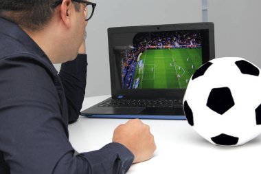 Latin yetişkin bir adam, iş saatlerinde futbol topunun yanında çalışırken laptopuyla Dünya Kupası maçı izliyor.
