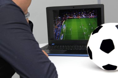Latin yetişkin bir adam, iş saatlerinde futbol topunun yanında çalışırken laptopuyla Dünya Kupası maçı izliyor.