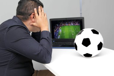 Latin yetişkin bir adam, iş saatlerinde futbol topunun yanında çalışırken laptopuyla Dünya Kupası maçı izliyor.