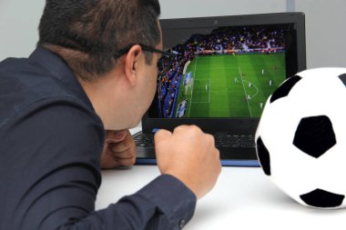 Latin yetişkin bir adam, iş saatlerinde futbol topunun yanında çalışırken laptopuyla Dünya Kupası maçı izliyor.