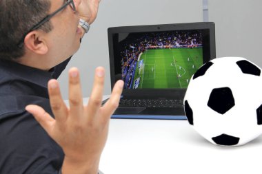Latin yetişkin bir adam, iş saatlerinde futbol topunun yanında çalışırken laptopuyla Dünya Kupası maçı izliyor.