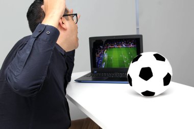 Latin yetişkin bir adam, iş saatlerinde futbol topunun yanında çalışırken laptopuyla Dünya Kupası maçı izliyor.