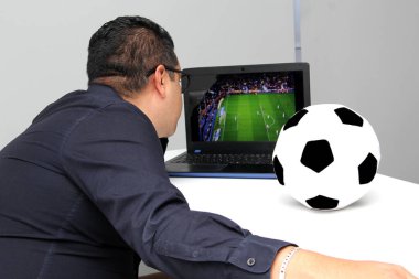 Latin yetişkin bir adam, iş saatlerinde futbol topunun yanında çalışırken laptopuyla Dünya Kupası maçı izliyor.