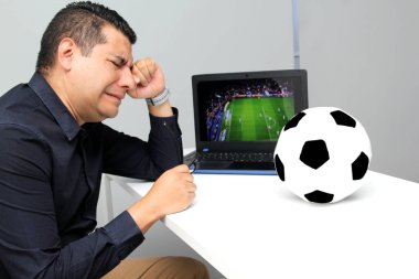 Latin yetişkin bir adam, iş saatlerinde futbol topunun yanında çalışırken laptopuyla Dünya Kupası maçı izliyor.