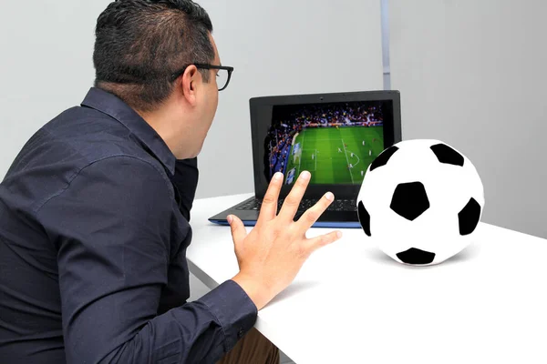 Latino adult man watches a World Cup soccer game on his laptop in his office while working next to a soccer ball during work hours in the morning