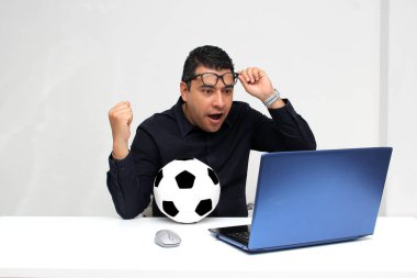 Latin yetişkin ofis çalışanı, iş saatleri boyunca futbol maçlarını bilgisayarından izliyor. Onu futbol topunun yanında heyecanlı, gergin ve şaşırmış görüyor.