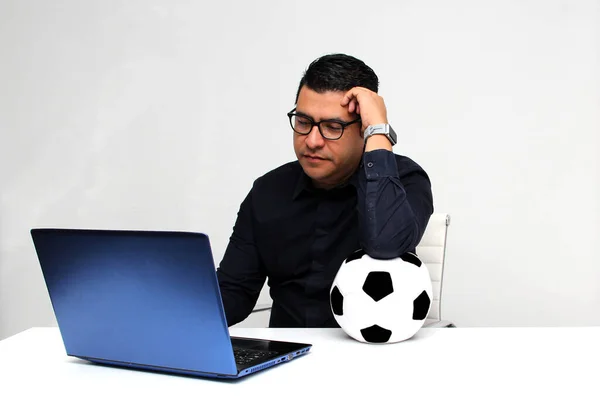 Latino adult office man watches football games on his work laptop during office hours in the morning, he sees him excited, nervous, surprised next to his soccer ball