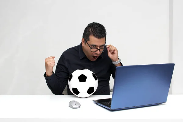 Latino adult office man watches football games on his work laptop during office hours in the morning, he sees him excited, nervous, surprised next to his soccer ball