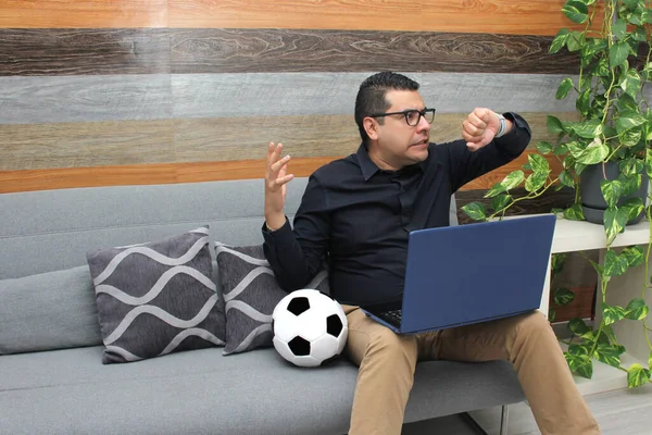 Latino adult office man watches football games on his work laptop during office hours in the morning, he sees him nervous, sad, stressed, angry next to his soccer ball