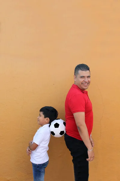 Latino dad and son share their love for soccer, they take a ball with their hands excited to watch the football game